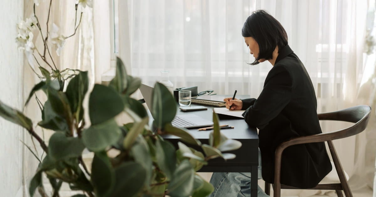 A business woman seated at a desk, busy with her business taxes. She's writing on a piece of paper in front of her laptop. A green plant is in the foreground.