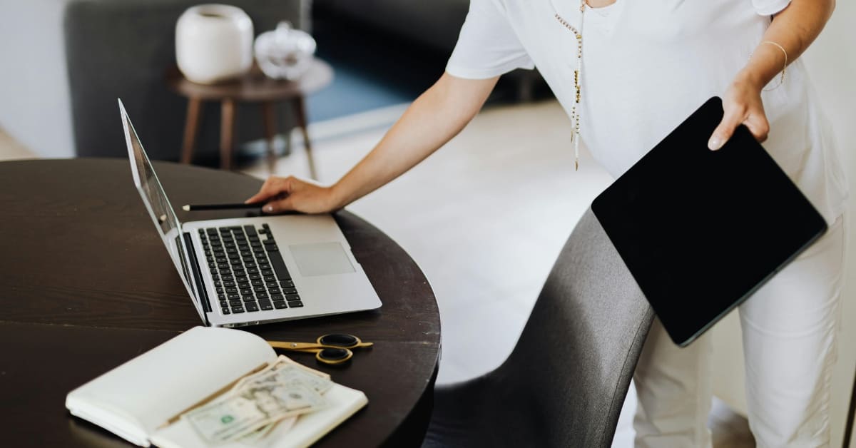 Person using a laptop and tablet at a desk with cash and a notebook, representing business finances and tax planning.