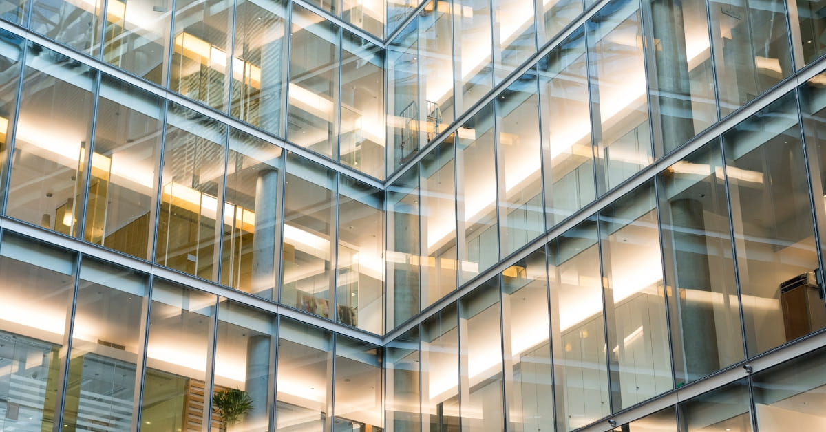 Modern office building interior with multiple levels of glass-walled offices, illuminated workspaces and geometric architectural lines.